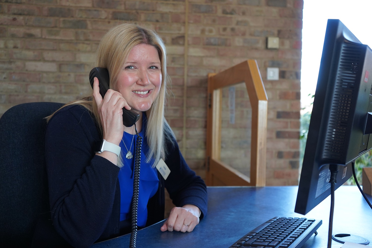 A woman holding a phone while sat at a desk with a computer, smiling at the camera.