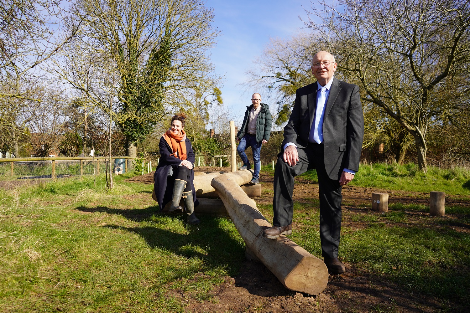 Three people stood and sat on some natural play logs in Rous Lench Community Garden.