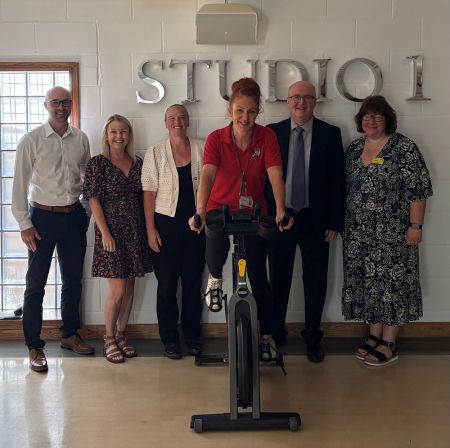 A woman on an exercise bike at the front of the picture with five people stood either side in the background in a line